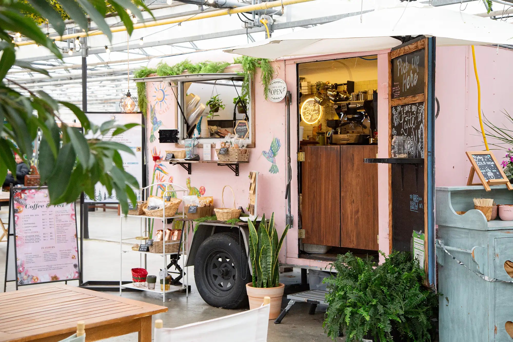 Pink coffee trailer with door and window open and lights inside, surrounded by plants inside a greenhouse