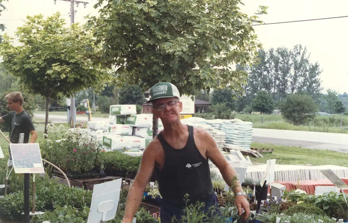 Adrian Vandermeer standing amongst tables of flowers in front yard, with skids of soil in background in roughly 1963