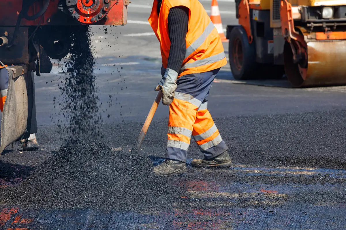 Person in high-visibility clothing shovelling asphalt with construction equipment in the background