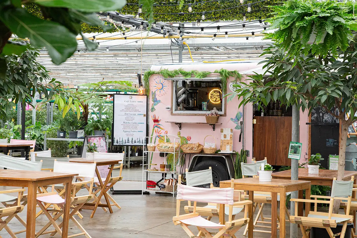 Pink coffee trailer with patio setup of wooden tables and chairs, surrounded by plants inside a greenhouse