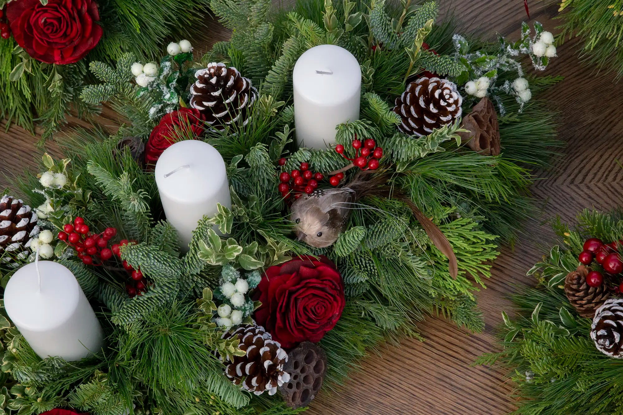 Evergreen centrepiece with three white pillar candles, pinecones, berries, red roses, and a faux brown bird.