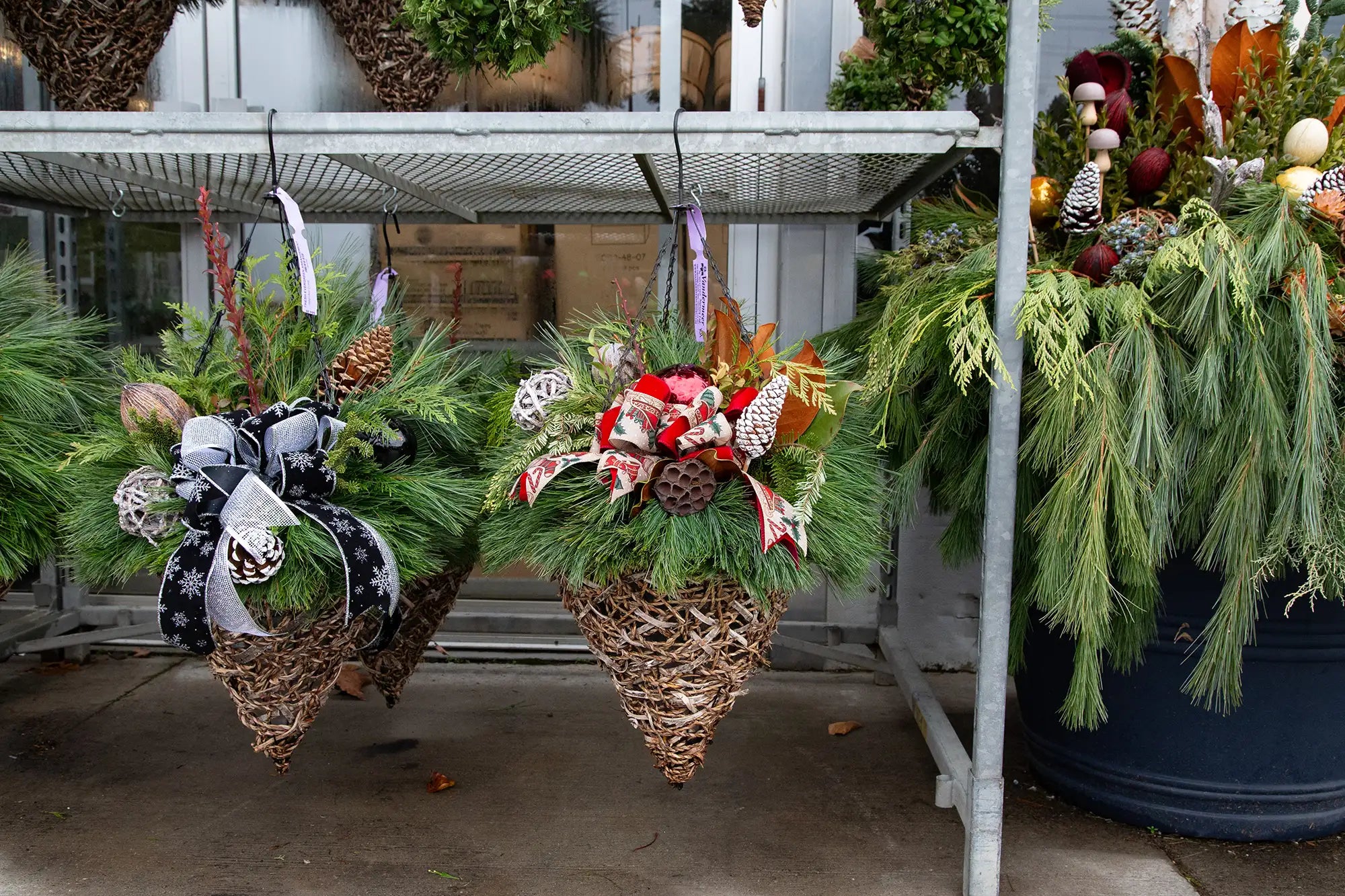 Decorative evergreen cone basket with greenery, ornaments, and bow hanging in an outdoor garden centre setting.