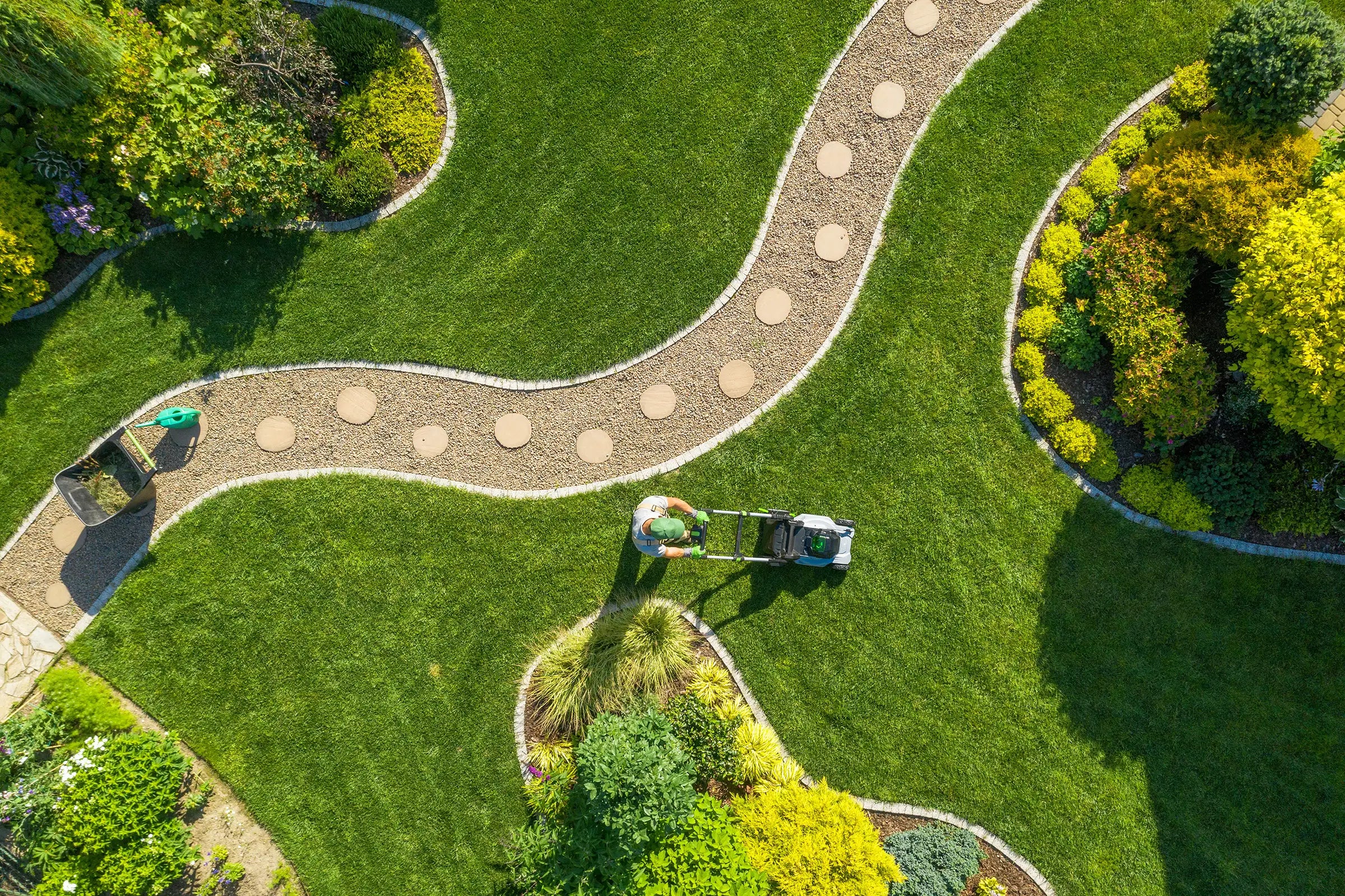 Bird's-eye view of a person mowing a lawn with a winding path and garden beds.