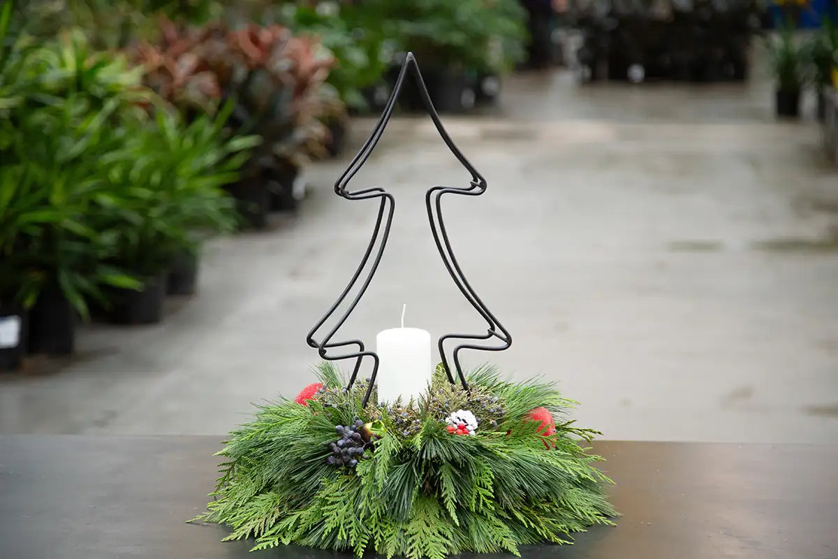 A festive holiday centerpiece featuring a black wire tree silhouette frame holding a white pillar candle, surrounded by a green pine wreath with small red berries and evergreen accents, set on a table in a plant nursery.