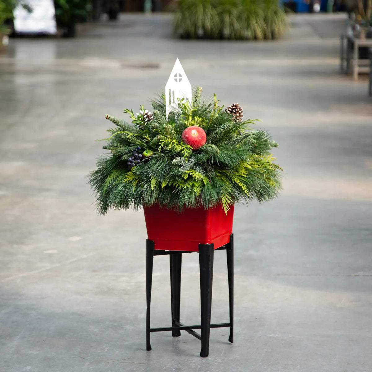 Decorative red Christmas pot filled with evergreen arrangement, decor, and small white house silhouette on a stand in an indoor setting.