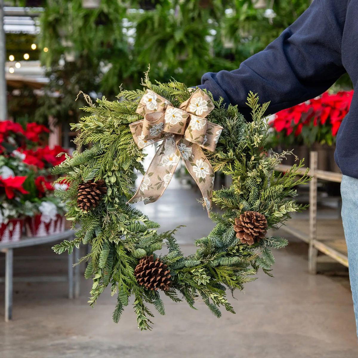 Person holding a Christmas wreath made of mixed evergreens with a decorative bow in a retail greenhouse setting.