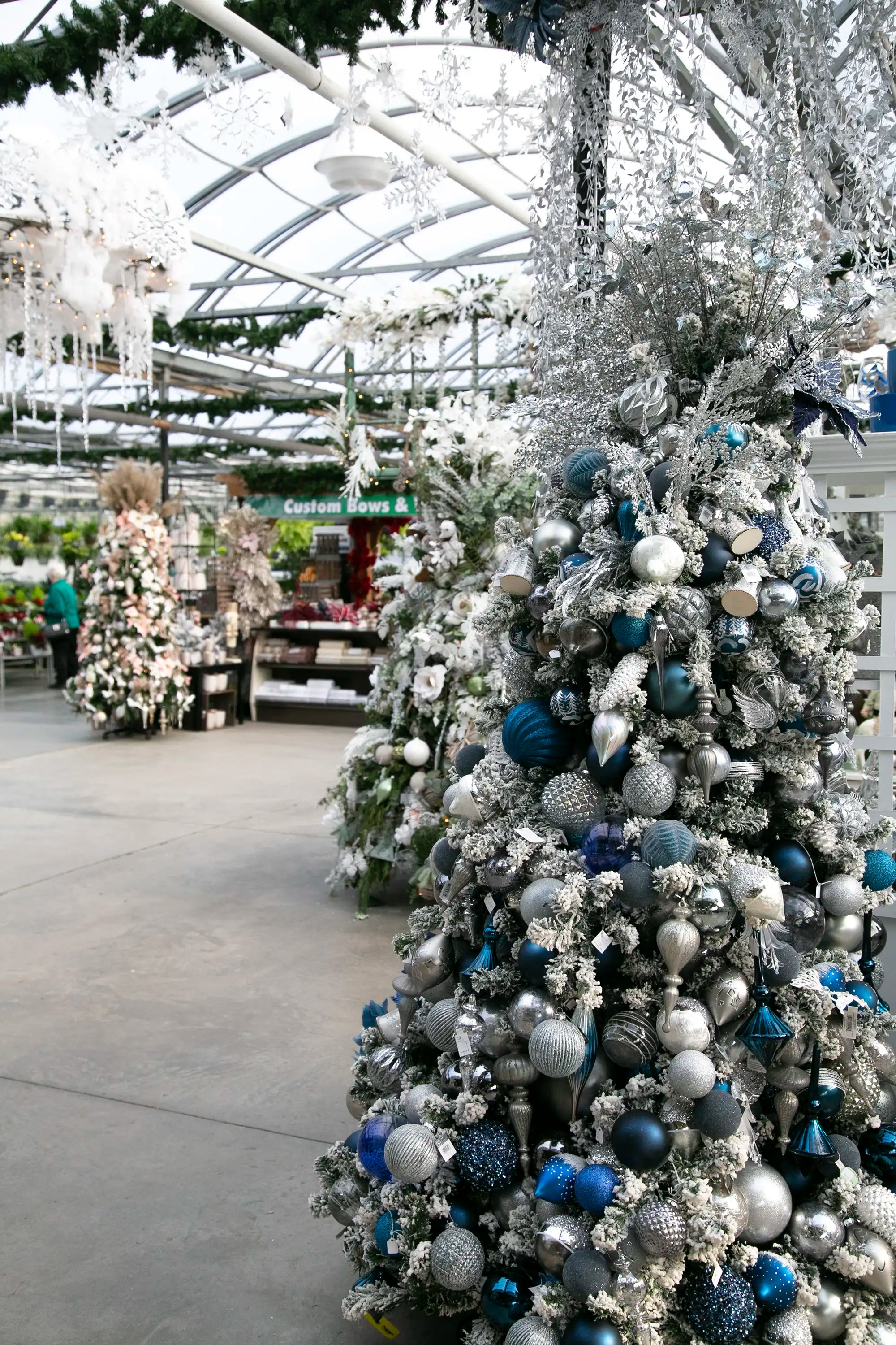 Designer-decorated Christmas tree with icy blue-themed ornaments in a festive greenhouse setting, and decorated trees in background