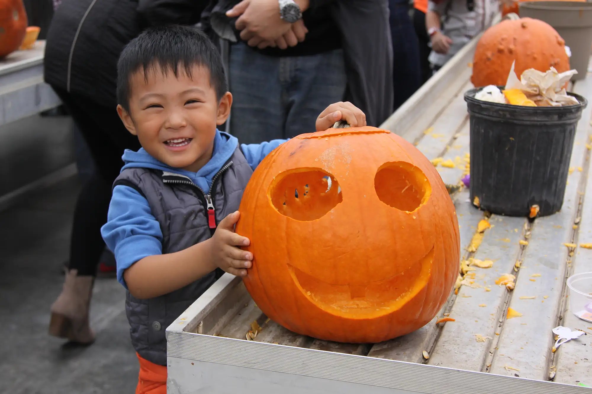 Child holding a carved pumpkin on a greenhouse table with people and pumpkins in the background.
