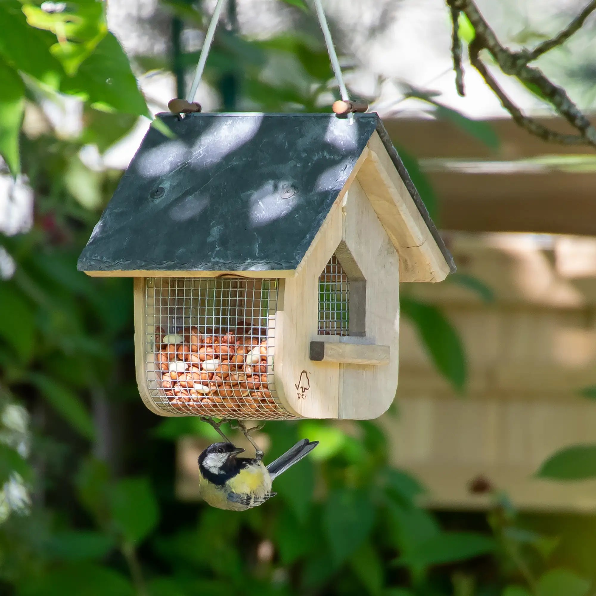 Wooden bird feeder with a bird perched on it, surrounded by green foliage.