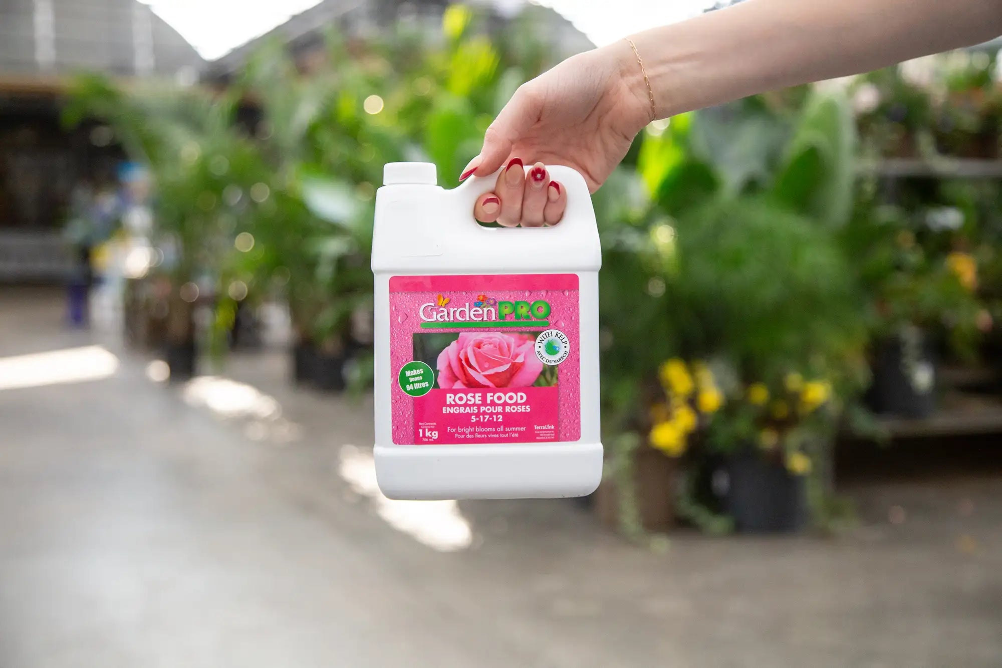 Hand holding a container of Garden Pro Rose Food in a greenhouse setting