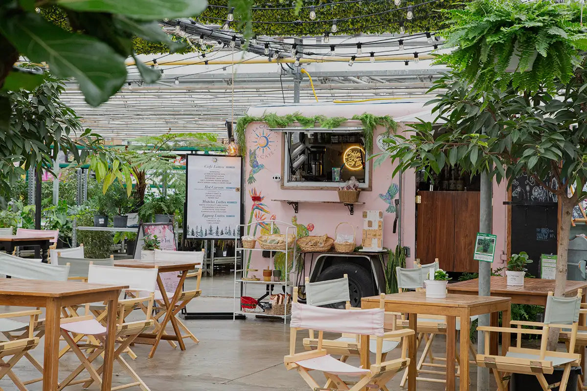 Pink coffee trailer with patio setup of wooden tables and chairs, surrounded by plants inside a greenhouse