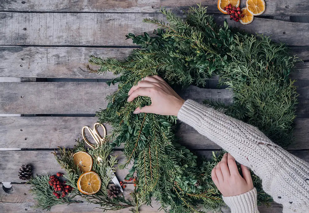 Person making wreath on wooden table with mixed evergreen boughs, pinecones, berries and orange slices.