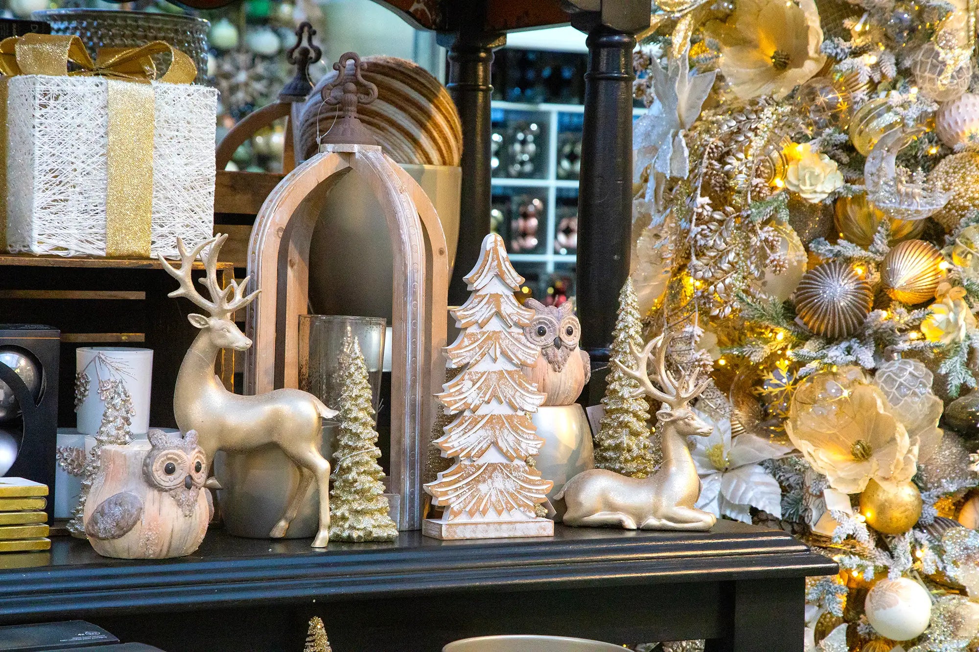Decorative items including a deer, owl, and Christmas tree figurines on a shelf with a Christmas tree in the background.