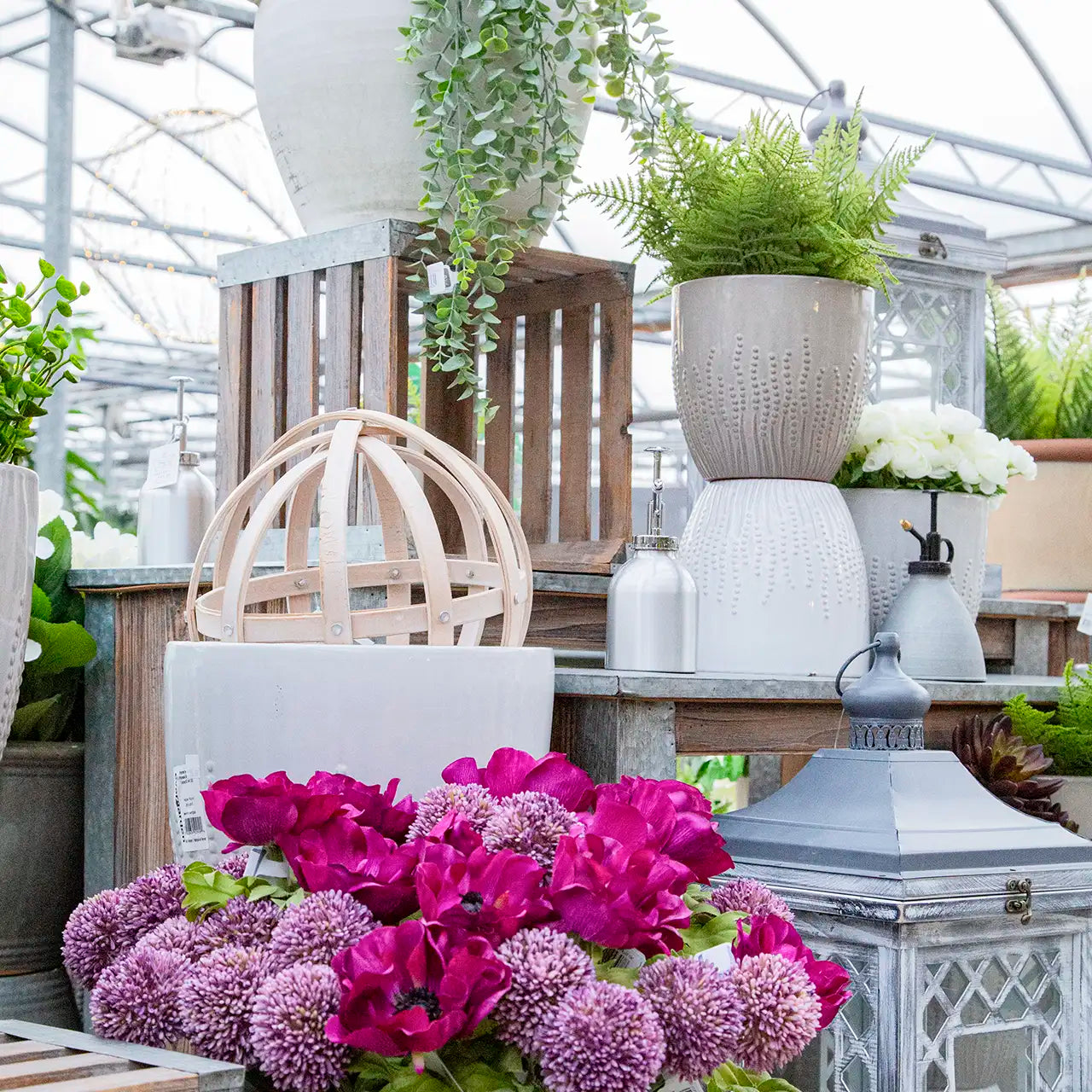 Faux pink florals and greenery in greenhouse display with cream ceramic pots and lanterns.