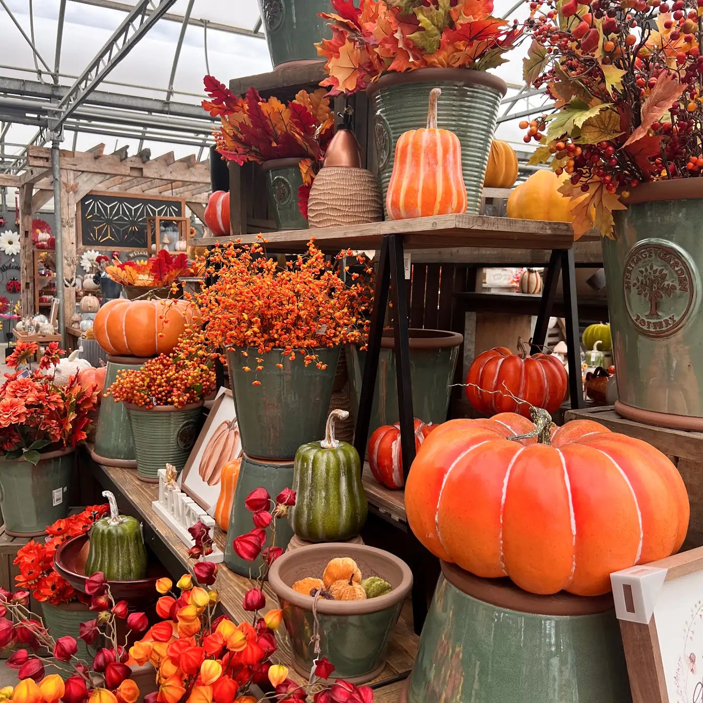 Decorative artificial pumpkins and fall-themed faux flowers, with muted teal ceramic pots in a greenhouse display