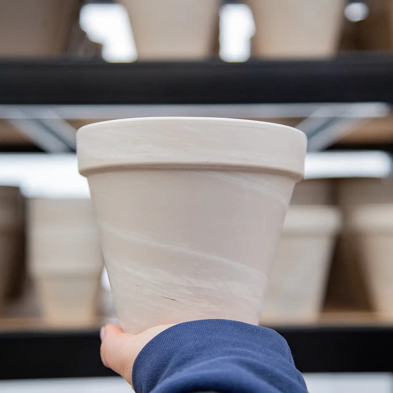 Person holding a light brown clay pot with shelves of pots in the background