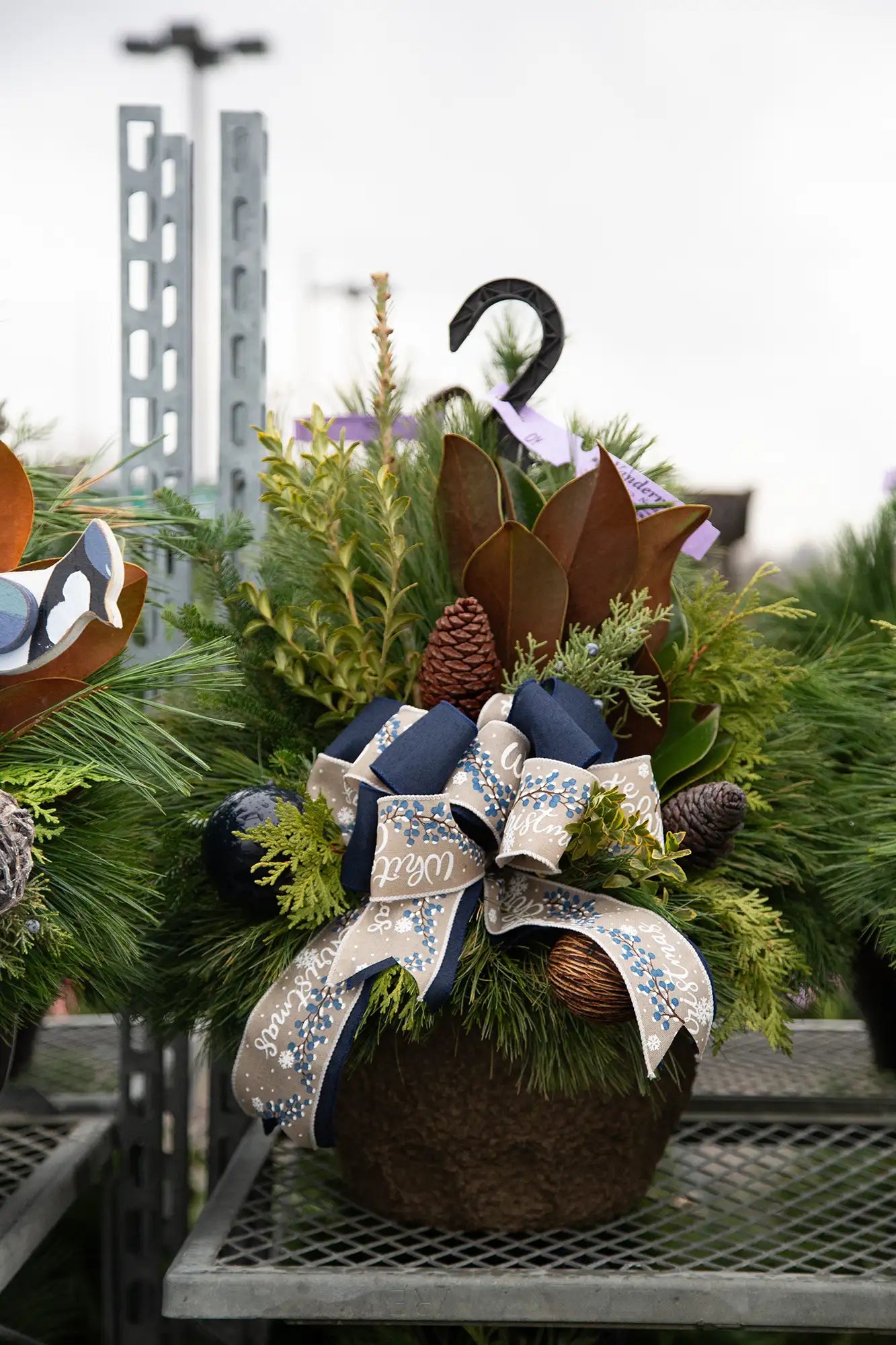 Decorative evergreen planter with greenery, ornaments, and blue and burlap bow in an outdoor garden centre setting.