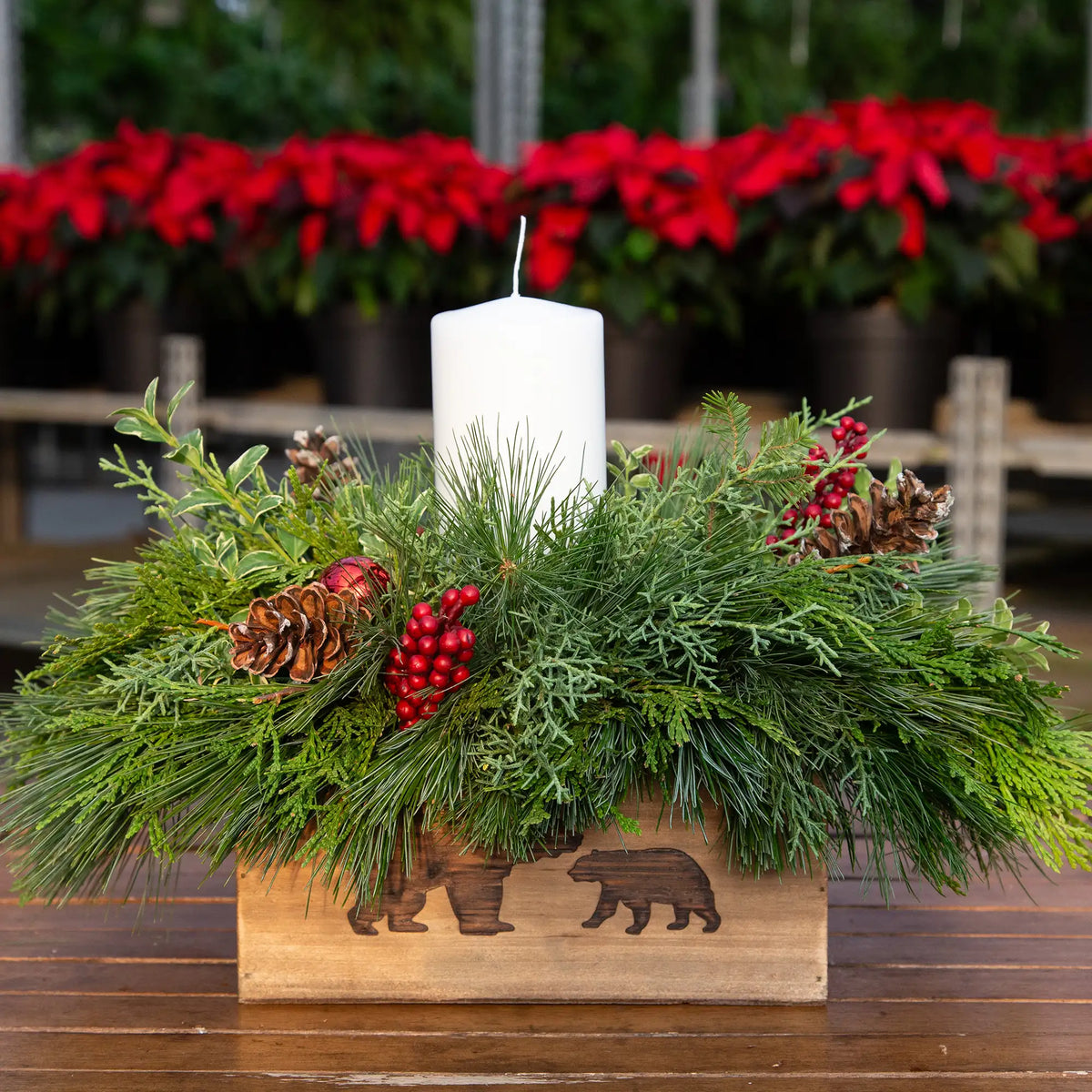 Decorative evergreen Christmas arrangement with a candle in a wooden box featuring bear illustrations, set against a blurred background of poinsettias.