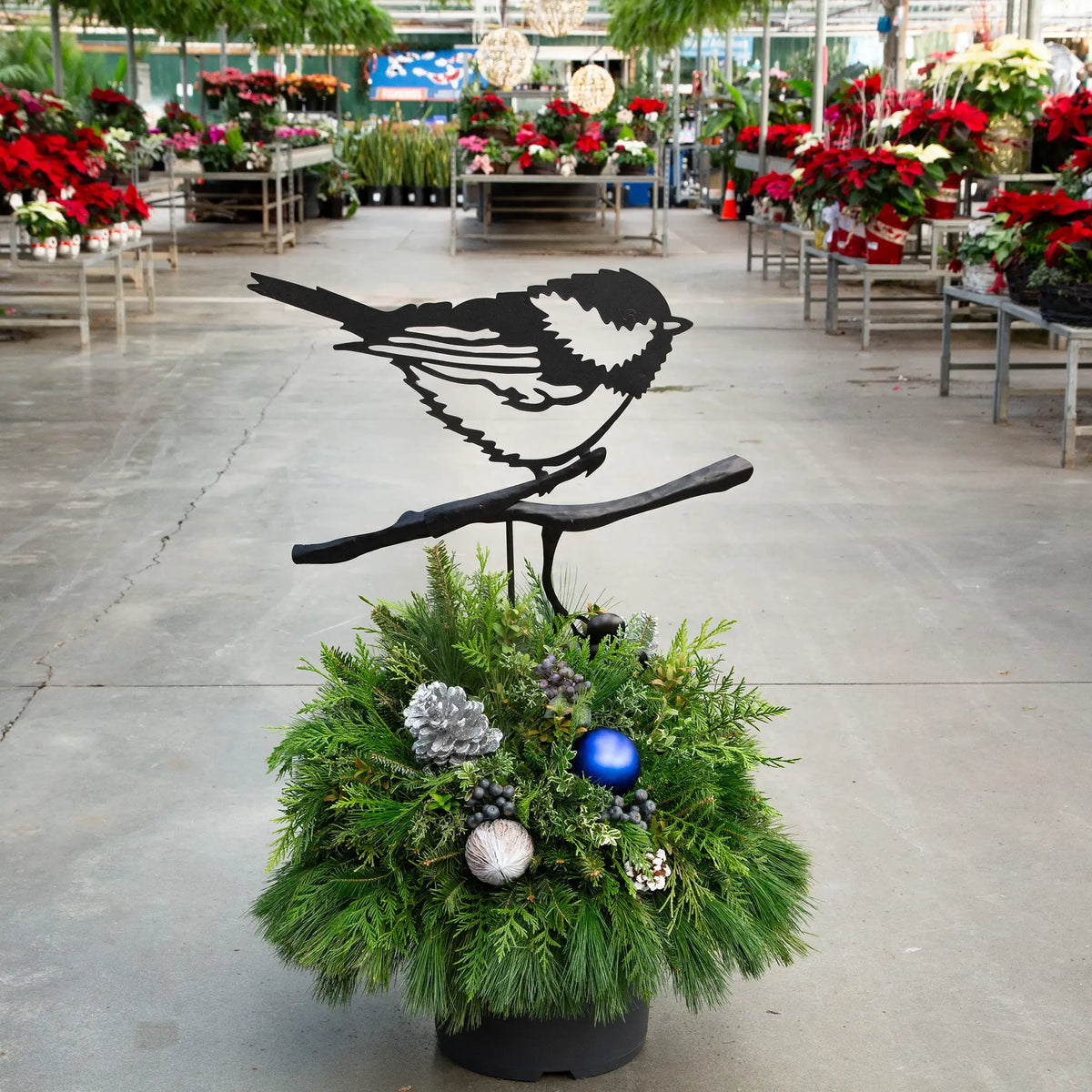 Evergreen arrangement with a black metal chickadee accent in it in a greenhouse setting.