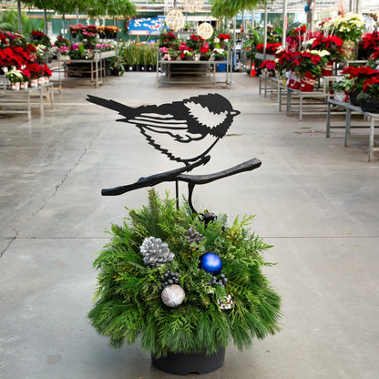 Evergreen arrangement with a black metal chickadee accent in it in a greenhouse setting.