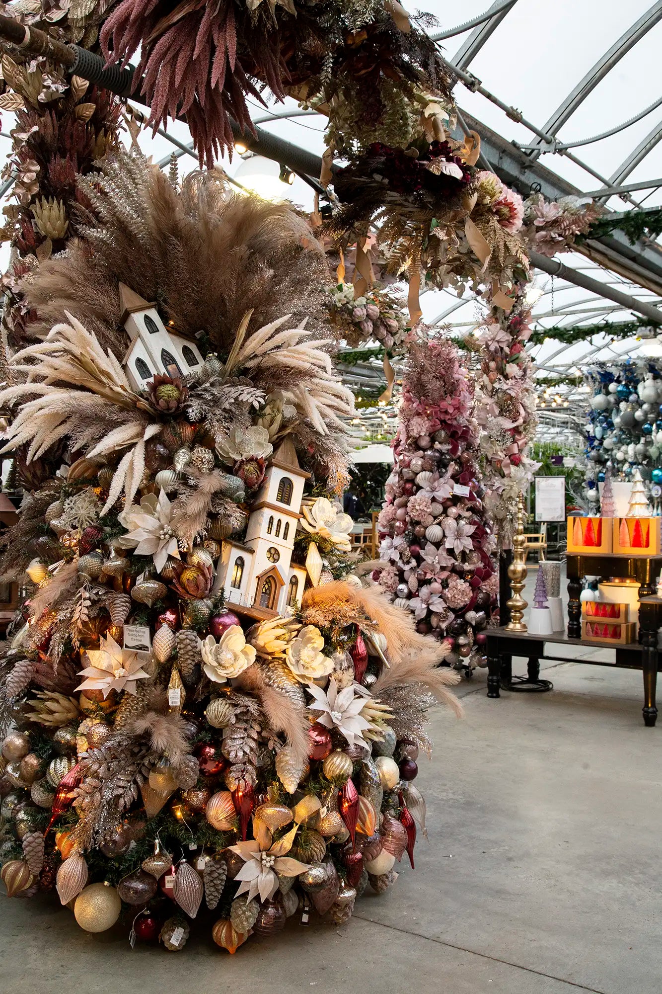Designer-decorated Christmas tree with themed ornaments in a festive greenhouse setting and a decorated pink tree in the background. 